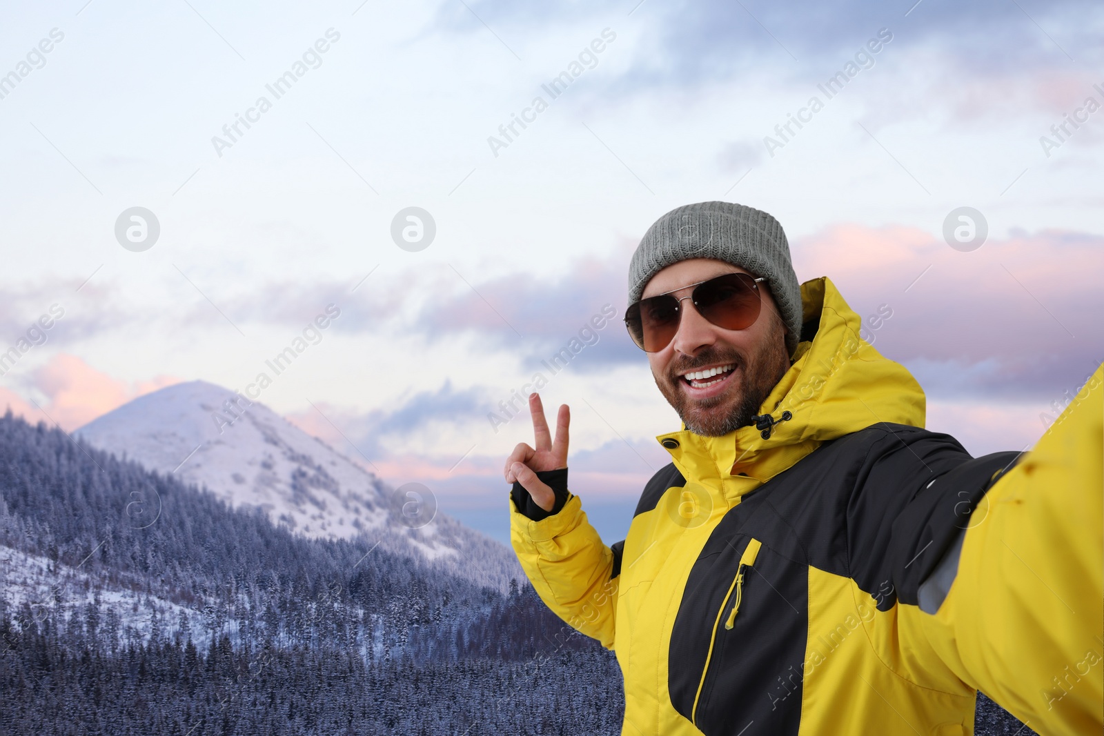 Image of Smiling man in sunglasses taking selfie and showing peace sign in mountains