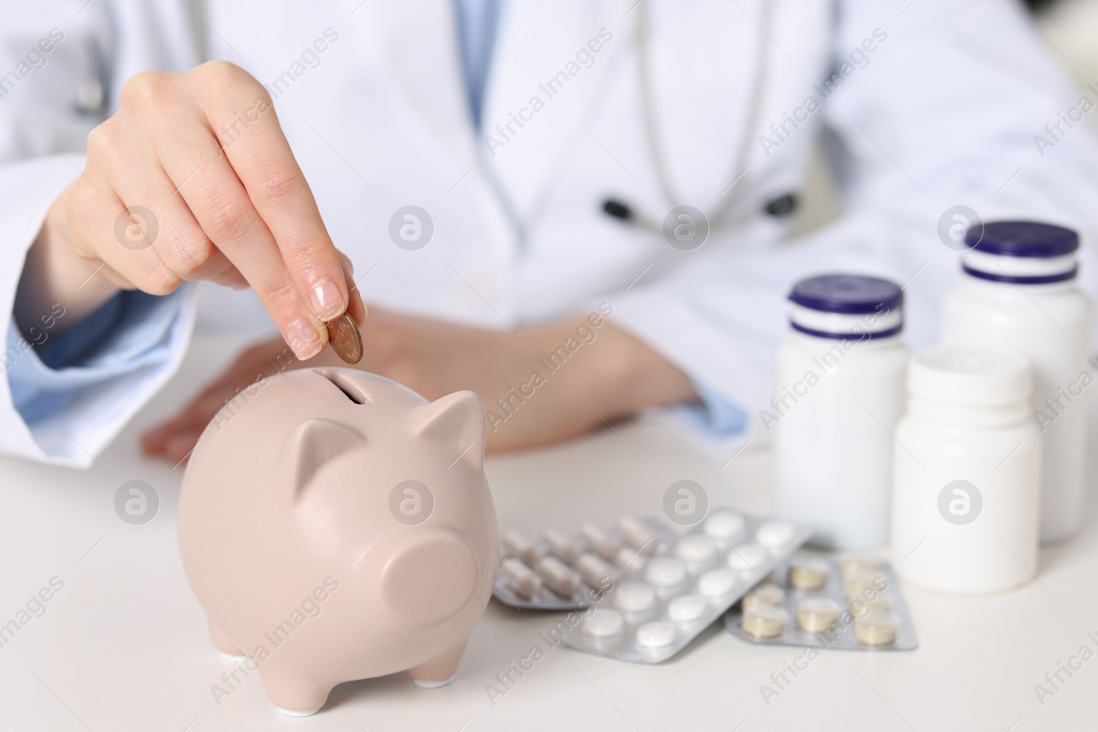 Photo of Doctor putting coin into piggy bank at white table, closeup