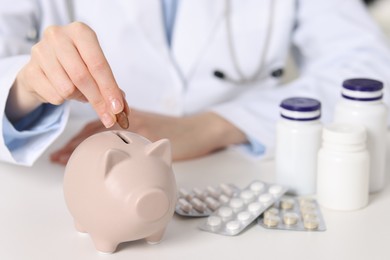 Photo of Doctor putting coin into piggy bank at white table, closeup