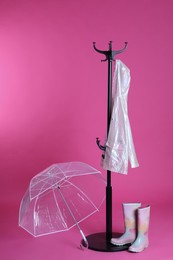 Photo of Open transparent umbrella, stylish rack with raincoat and rubber boots on pink background