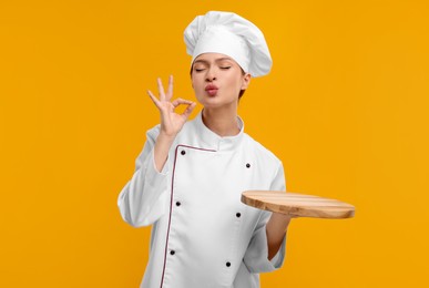 Young woman chef in uniform holding empty wooden board and showing perfect sign on orange background