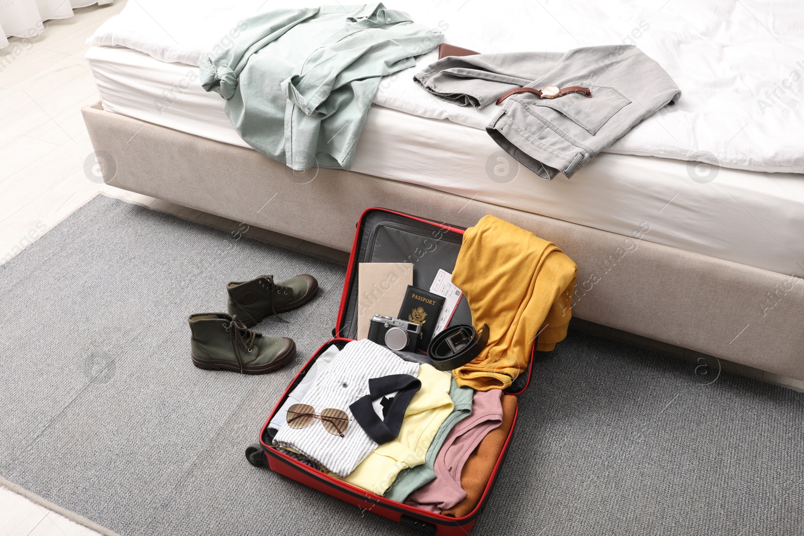 Photo of Open suitcase with clothes, accessories and shoes on floor near bed indoors