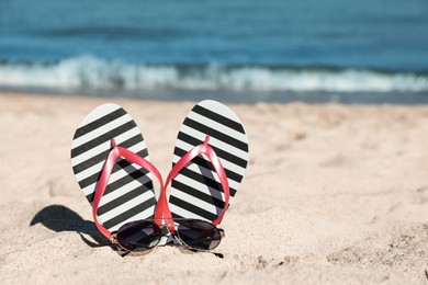 Photo of Striped flip flops and sunglasses on sandy beach near sea. Space for text