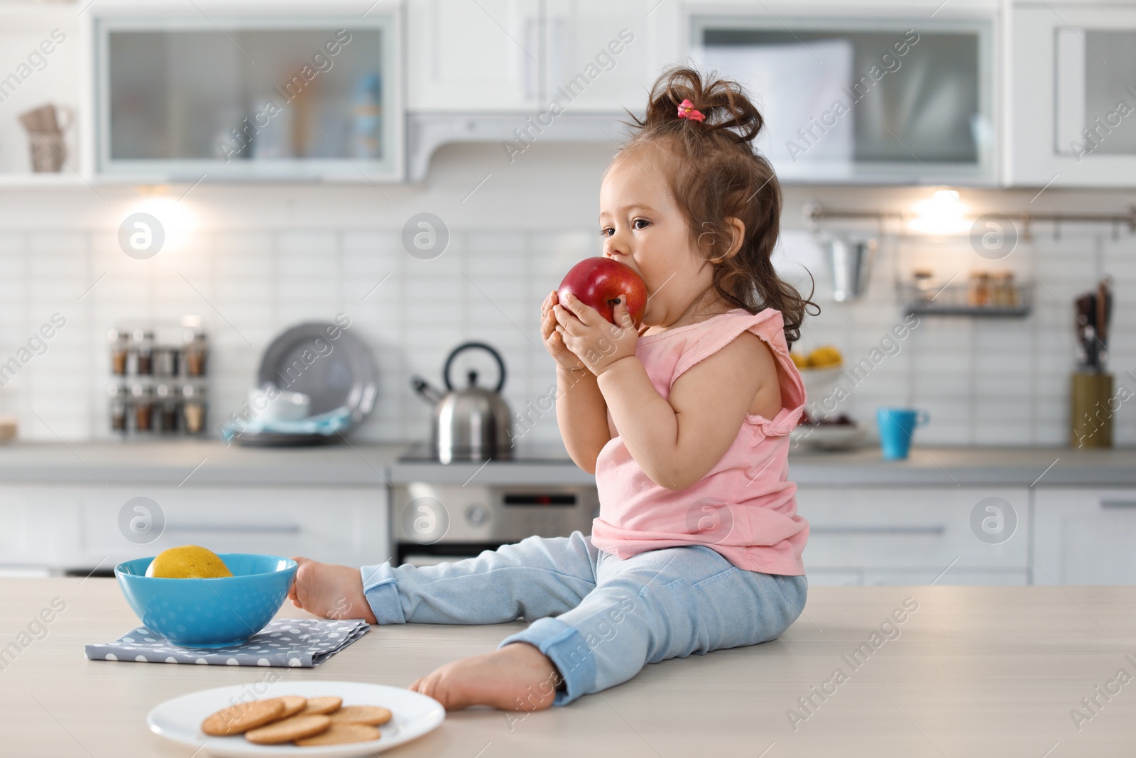 Photo of Adorable little baby girl eating apple on table in kitchen