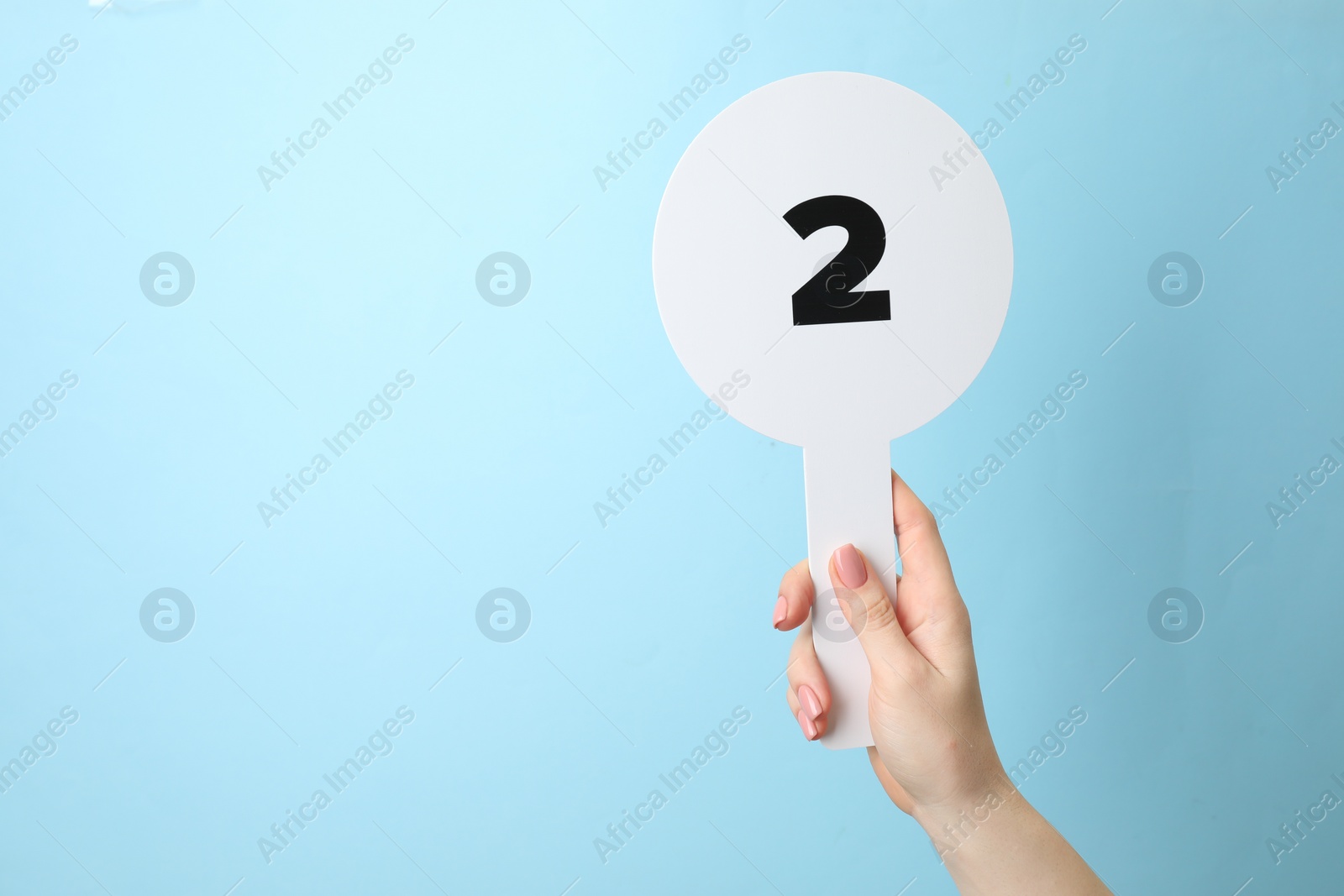 Photo of Woman holding auction paddle with number 2 on light blue background, closeup. Space for text