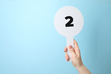 Woman holding auction paddle with number 2 on light blue background, closeup. Space for text