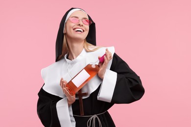 Happy woman in nun habit and sunglasses holding bottle of wine on pink background. Space for text