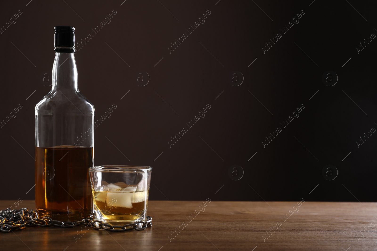 Photo of Alcohol addiction. Chain near bottle and glass of whiskey on wooden table. Space for text