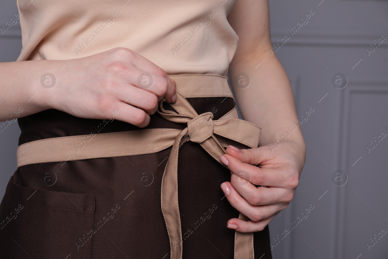 Photo of Woman putting on brown apron against grey wall, closeup