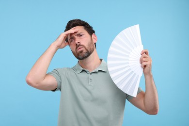 Unhappy man with hand fan suffering from heat on light blue background