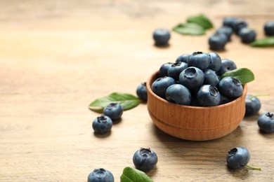 Photo of Bowl of tasty fresh blueberries with leaves on wooden table, closeup. Space for text