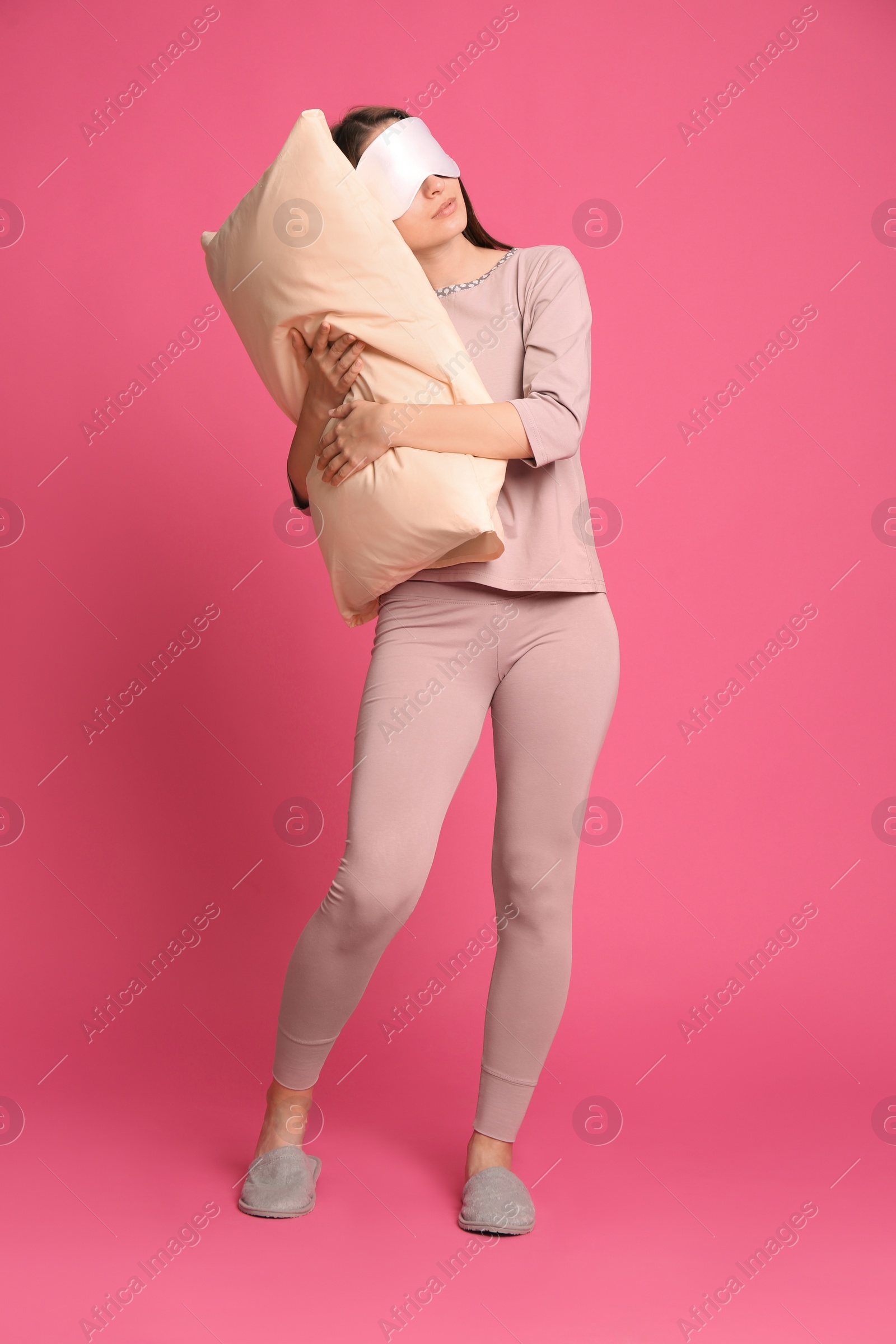 Photo of Young woman wearing pajamas, mask and slippers with pillow in sleepwalking state on pink background