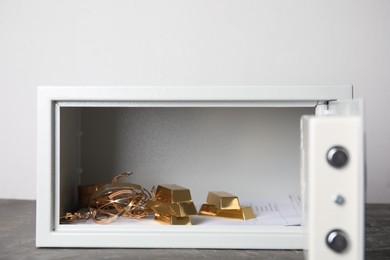 Open steel safe with gold bars and jewelry on grey table against light background