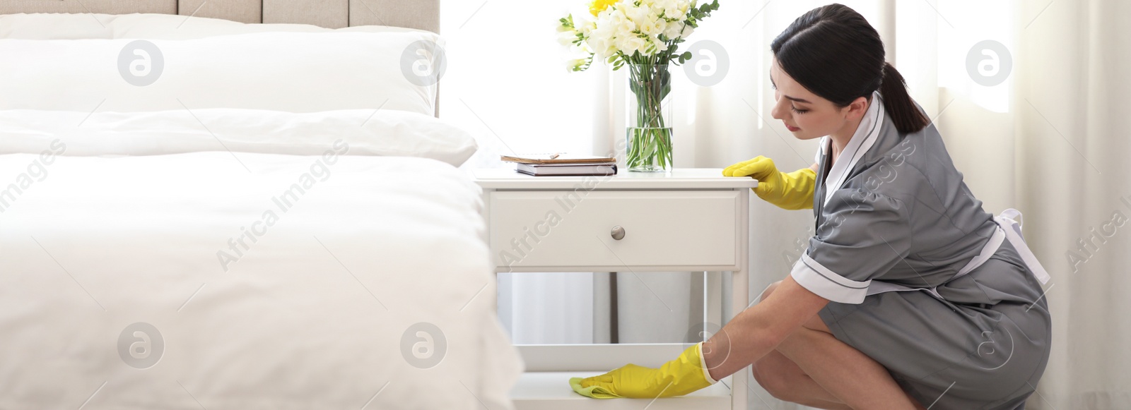 Image of Young chambermaid wiping dust from nightstand in bedroom. Banner design