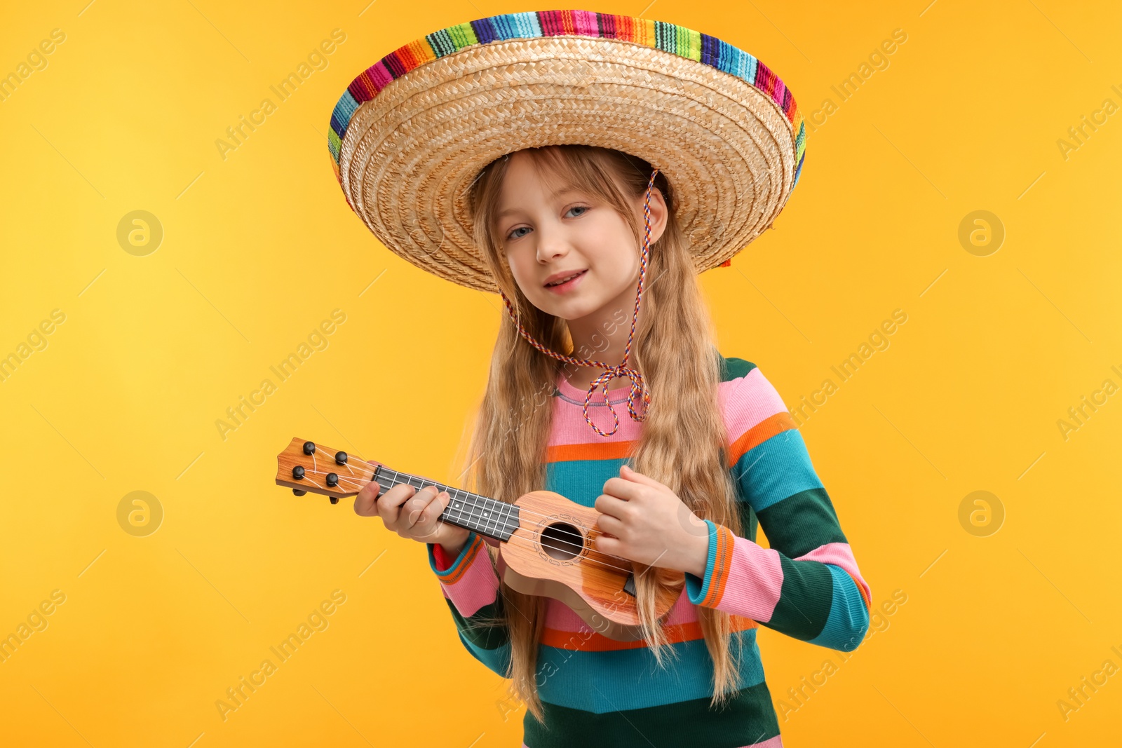 Photo of Cute girl in Mexican sombrero hat playing ukulele on orange background