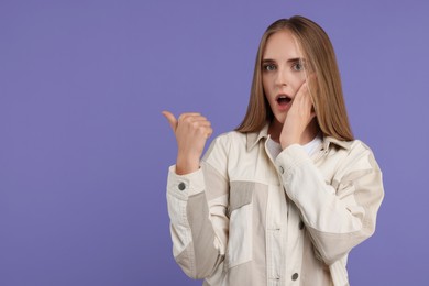 Embarrassed young woman pointing at something on violet background, space for text