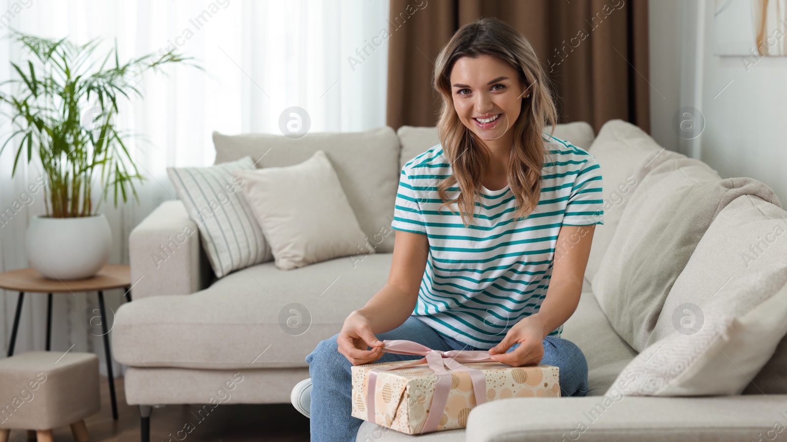 Photo of Beautiful young woman wrapping gift on sofa at home