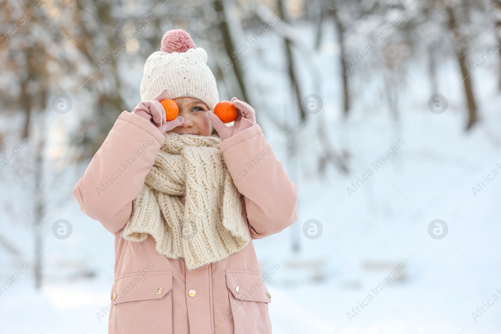 Photo of Cute little girl covering eye with tangerines in snowy park on winter day, space for text