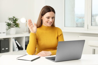 Photo of Woman waving hello during video chat via laptop at table in office