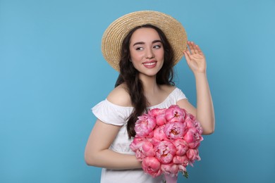 Photo of Beautiful young woman in straw hat with bouquet of pink peonies against light blue background