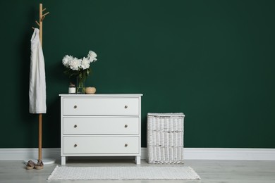 Photo of Modern white chest of drawers, coat stand and wicker basket near green wall indoors. Space for text