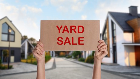 Image of Woman holding sign with text GARAGE SALE and blurred view of modern houses