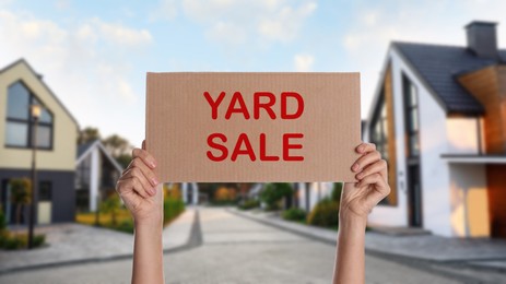 Woman holding sign with text GARAGE SALE and blurred view of modern houses