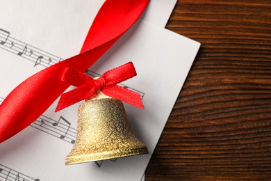 Photo of Golden shiny bell with red bow and music sheet on wooden table, flat lay. Space for text. Christmas decoration
