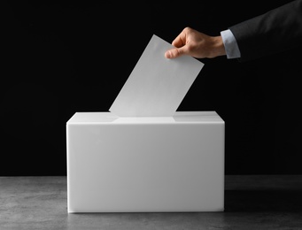 Man putting his vote into ballot box on table against black background, closeup