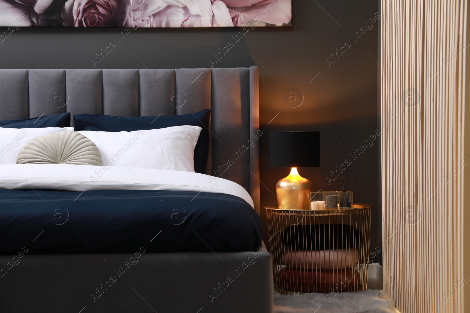 Photo of Comfortable bed with cushions, lamp and different decor on bedside table in room. Stylish interior