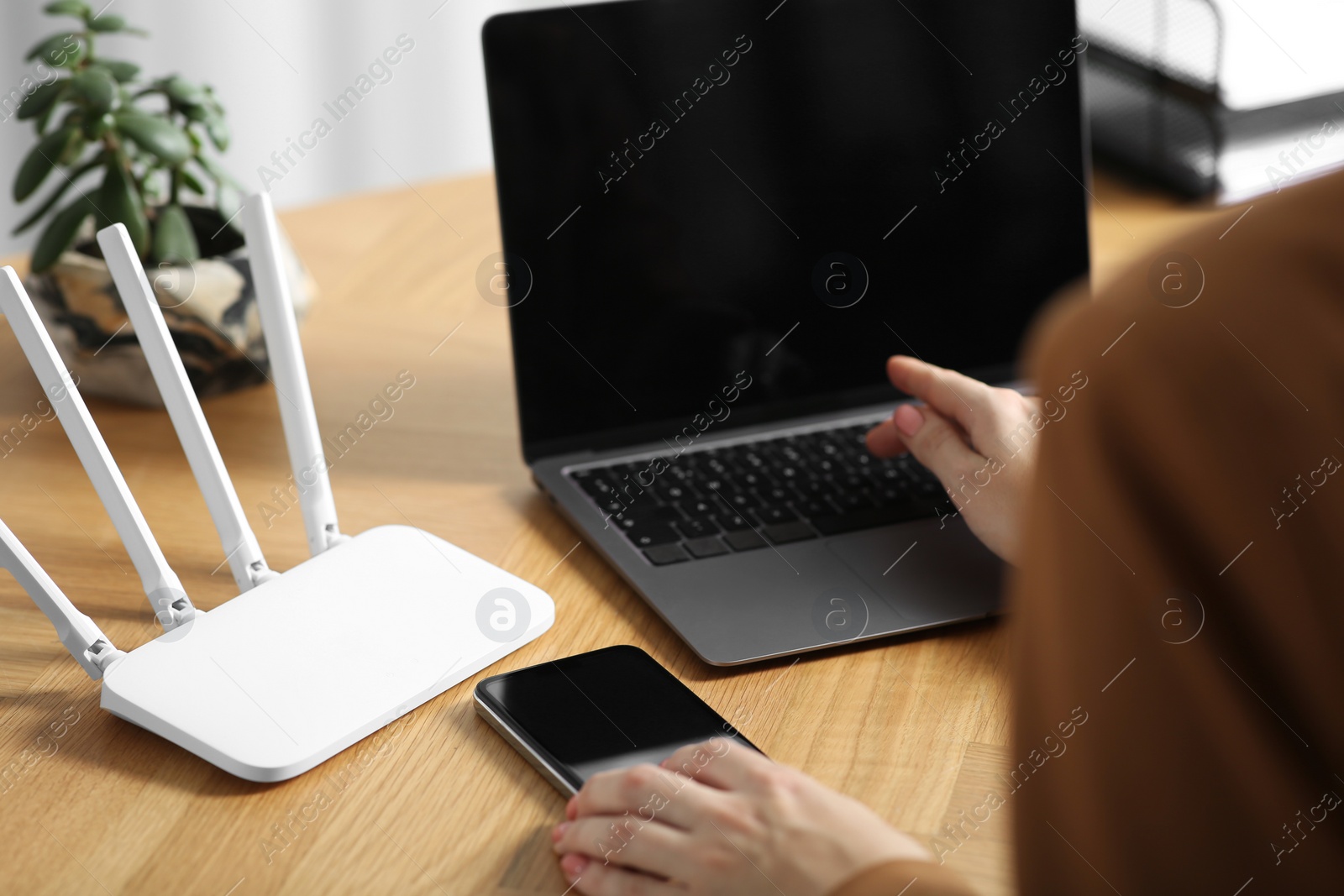 Photo of Woman with smartphone and laptop connecting to internet via Wi-Fi router at table indoors, closeup