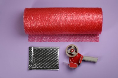 Photo of Bubble wrap roll, padded envelope and tape dispenser on lilac background, flat lay