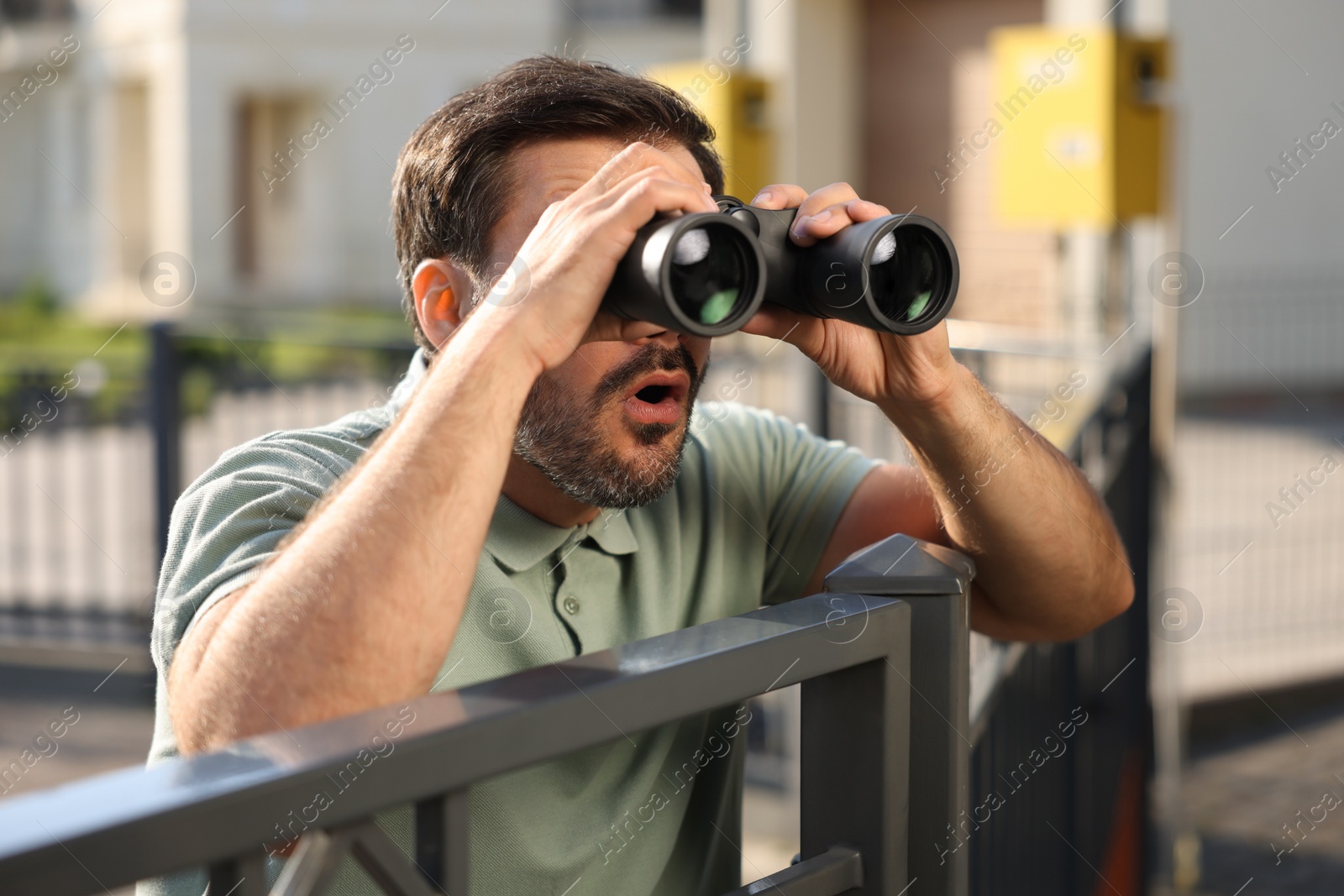 Photo of Concept of private life. Curious man with binoculars spying on neighbours over fence outdoors