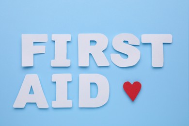 Photo of Words First Aid made of white letters and cardboard heart on light blue background, flat lay
