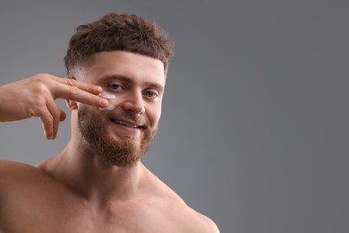 Handsome man applying moisturizing cream onto his face on grey background, space for text