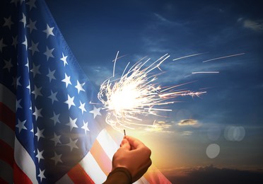 Image of 4th of July - Independence Day of USA. Woman holding burning sparkler near American flag outdoors at sunset, closeup