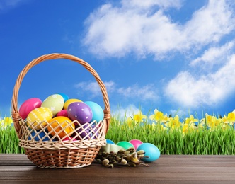 Image of Colorful Easter eggs in wicker basket and willow branches on wooden table outdoors, space for text