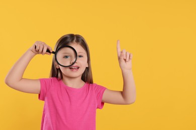 Photo of Cute little girl looking through magnifier glass and pointing on yellow background. Space for text