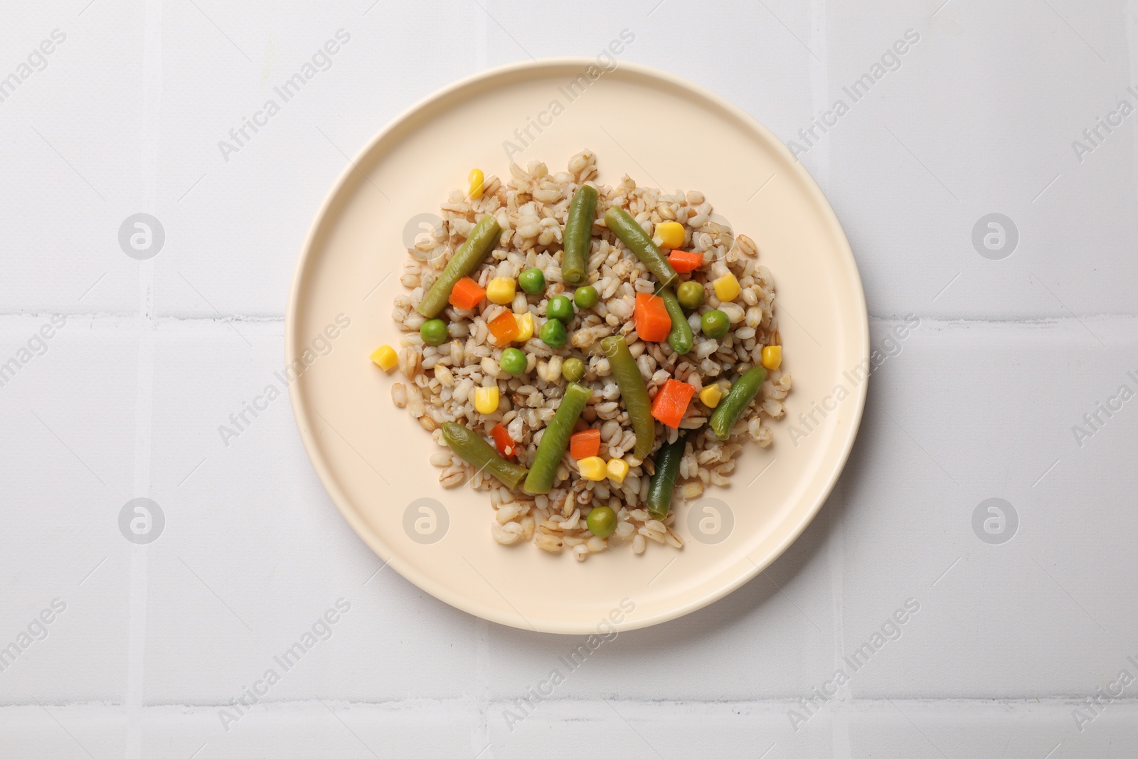 Photo of Delicious pearl barley with vegetables on white tiled table, top view