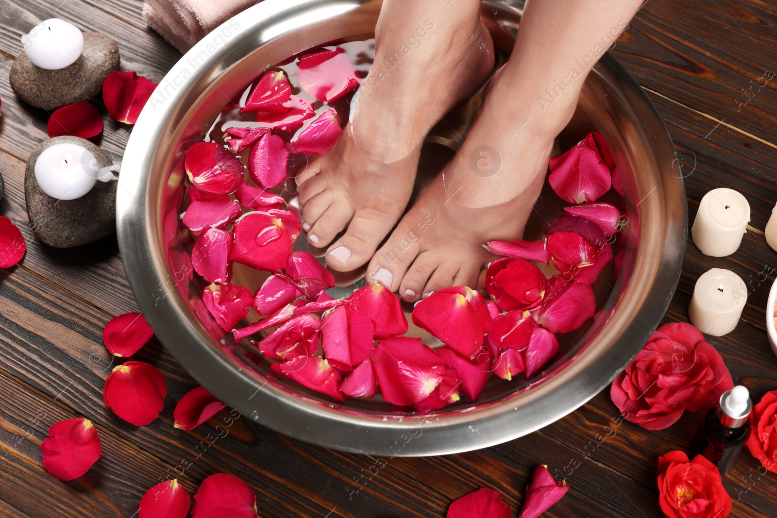 Photo of Woman soaking her feet in bowl with water and red rose petals on wooden floor, closeup. Pedicure procedure