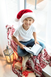 Photo of Cute little child in Santa hat reading Christmas story while sitting on windowsill at home