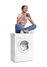 Beautiful woman with cup of drink sitting on washing machine against white background