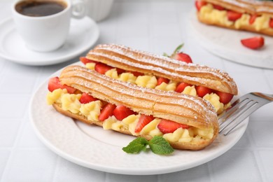 Photo of Delicious eclairs filled with cream, strawberries and mint on white tiled table