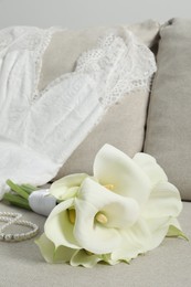 Photo of Beautiful calla lily flowers tied with ribbon, wedding dress and necklace on sofa