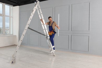 Electrician in uniform with ceiling lamp on metal ladder indoors