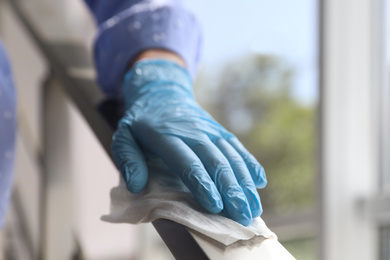 Photo of Woman in latex gloves cleaning railing with wet wipe indoors, closeup