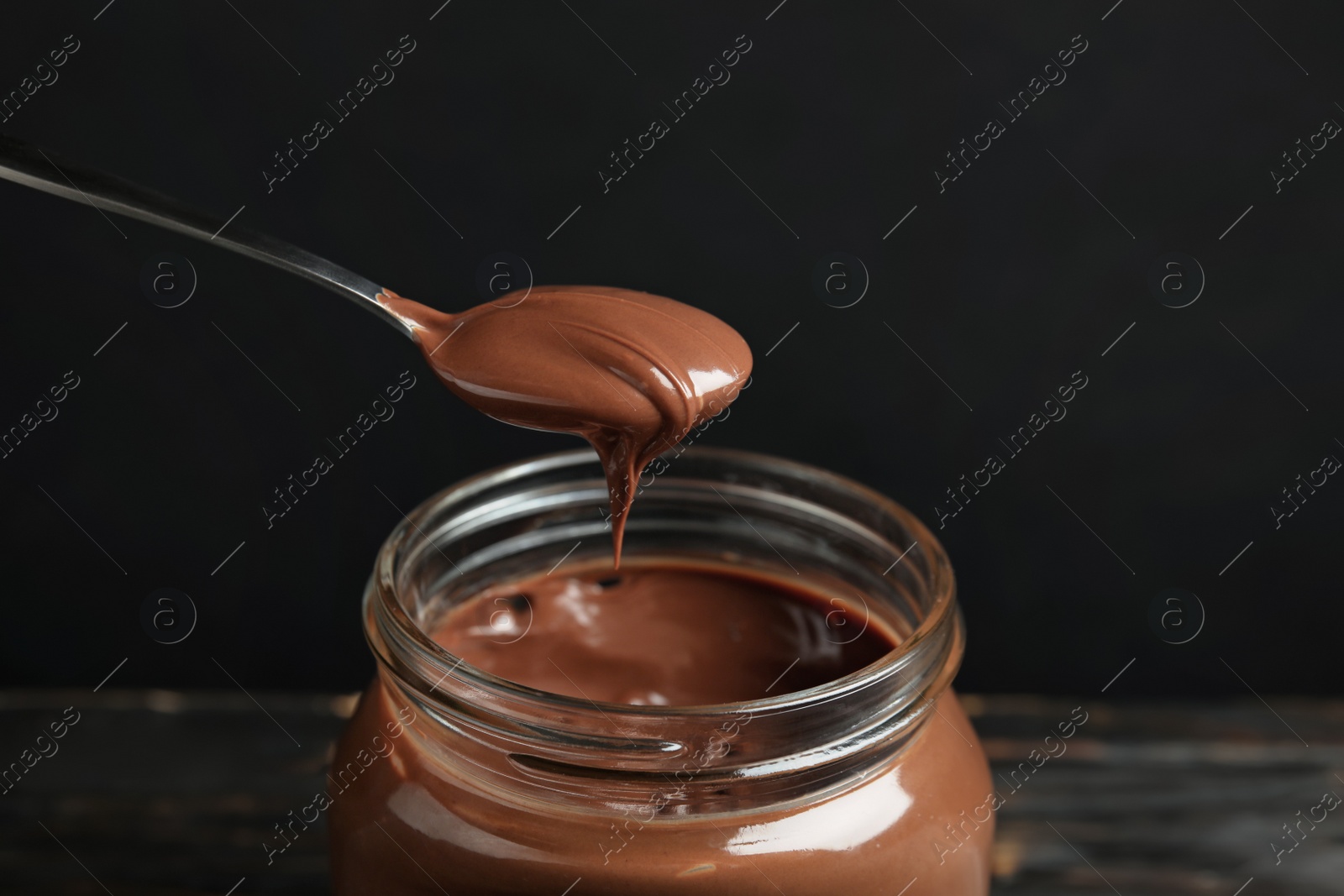 Photo of Spoon with tasty chocolate cream over glass jar on wooden table against dark background