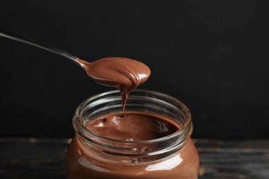 Spoon with tasty chocolate cream over glass jar on wooden table against dark background