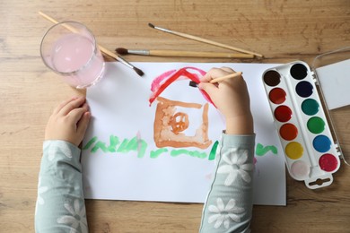 Cute little girl drawing house with brush at wooden table, top view. Child`s art