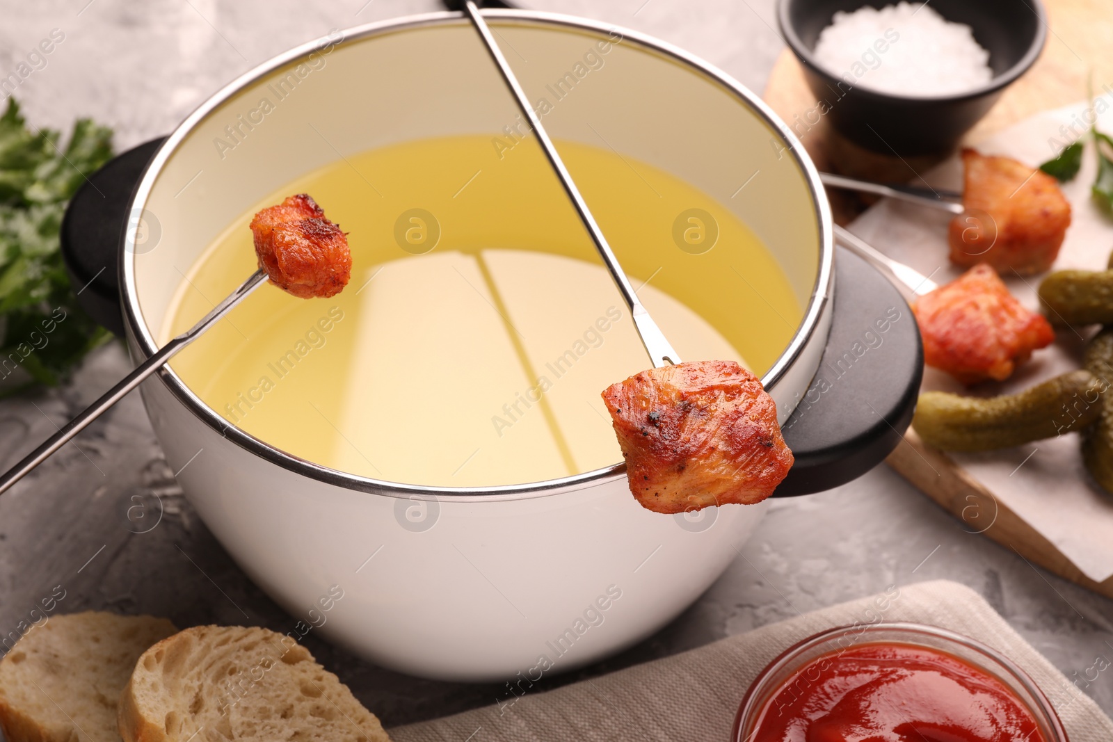 Photo of Fondue pot, forks with fried meat pieces and other products on grey textured table, closeup